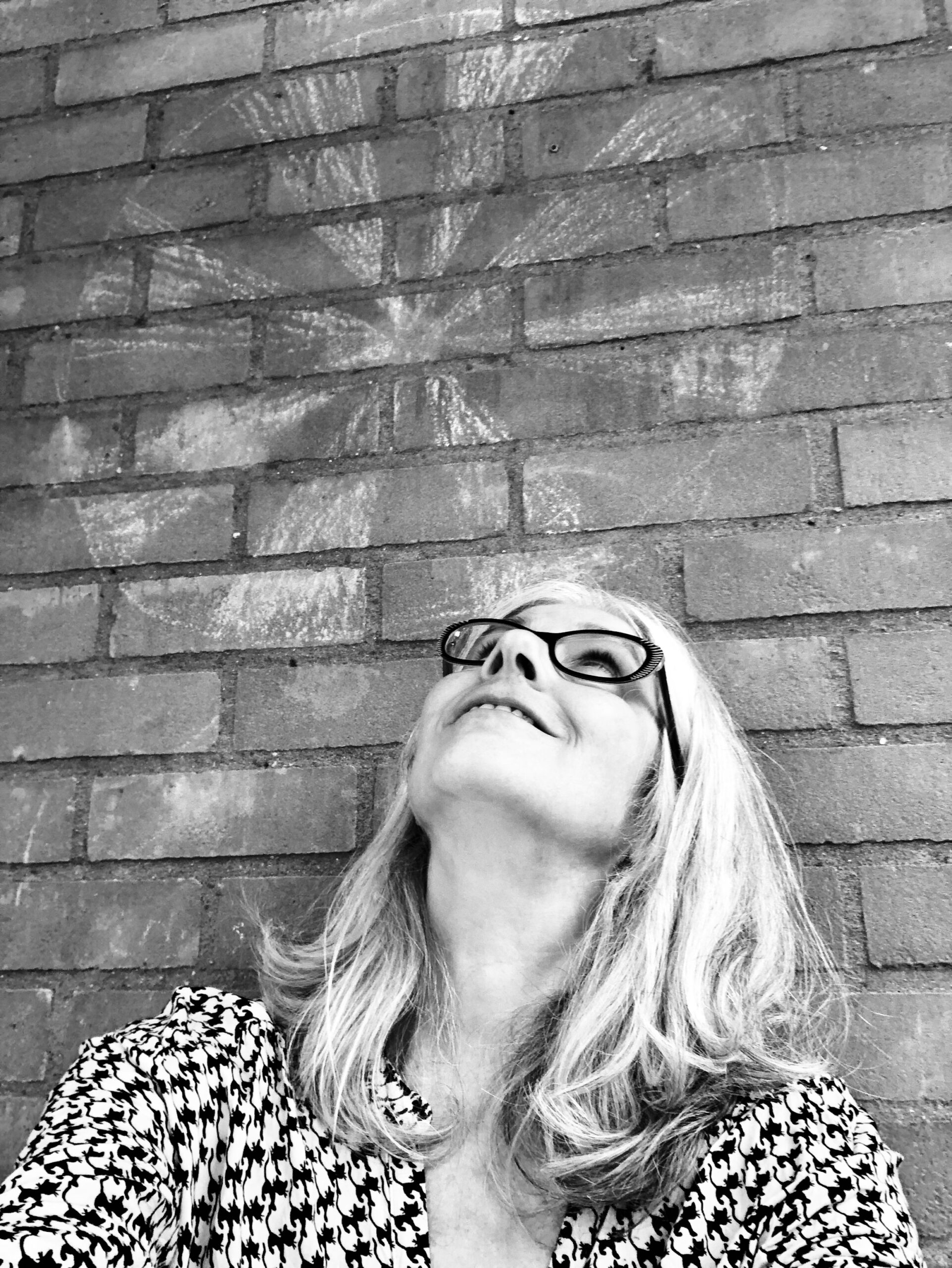 Zwart-wit portret van Reggie Voigtländer, staand bij een muur met een kindertekening van een windmolen, terwijl ze omhoog kijkt.