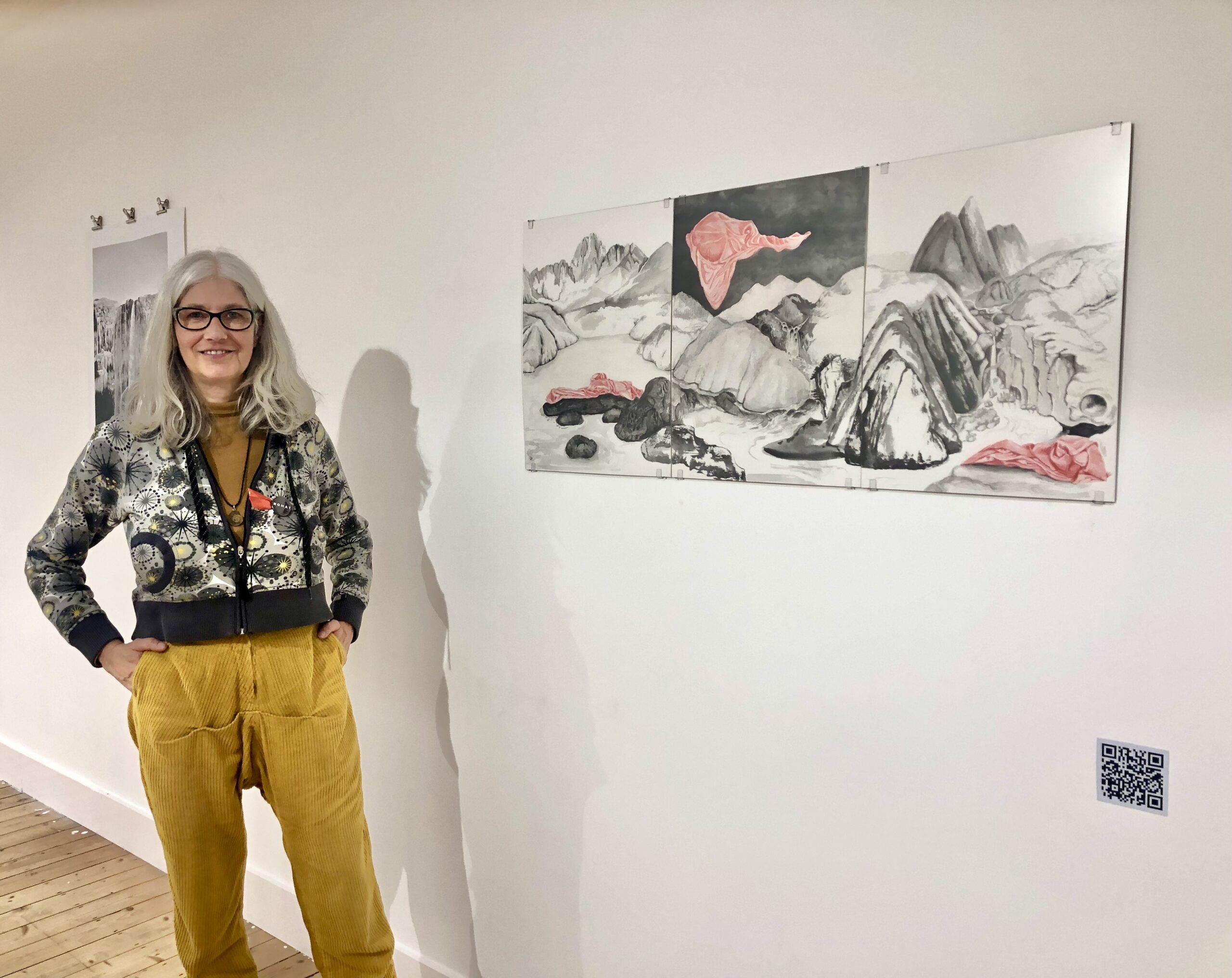 Reggie Voigtländer, kunstenaar en coach, staand in een galerie tijdens een expositie, met haar eigen kunstwerken zichtbaar op de achtergrond.