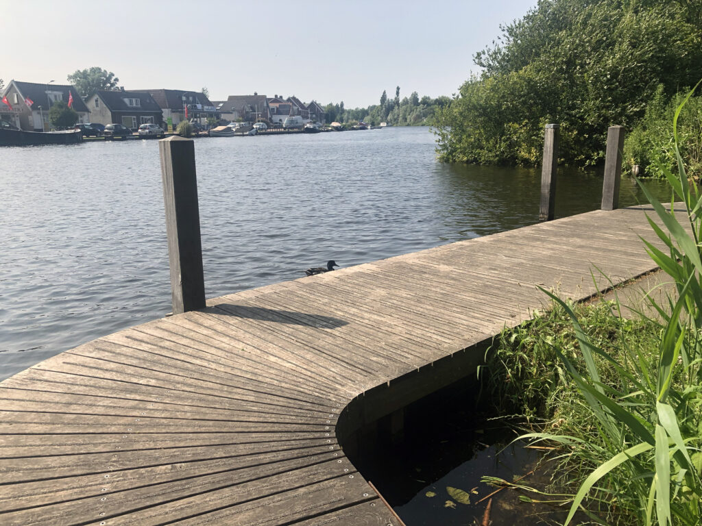 Vlonder langs een kanaal dat naar rechts afbuigt, met een heiige achtergrond van huizen aan de overkant.